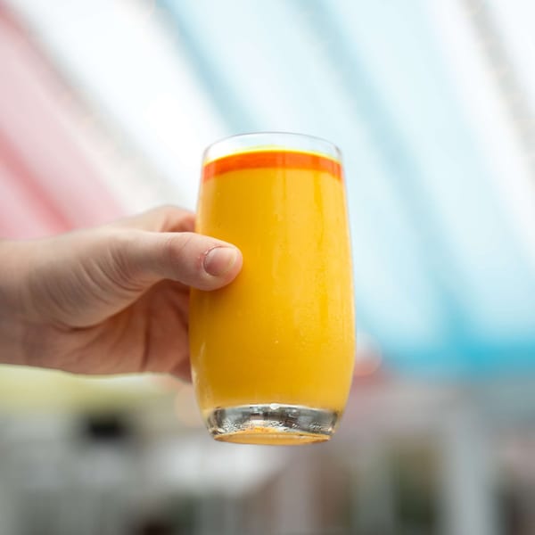A hand holds a tall glass of vibrant orange mango lassi, with a colorful background.