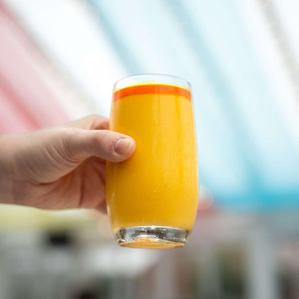 A hand holds a tall glass of vibrant orange mango lassi, with a colorful background.