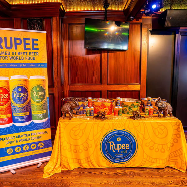 Rupee beer cans and buckets on a yellow tablecloth with elephant statues, next to a promotional banner.