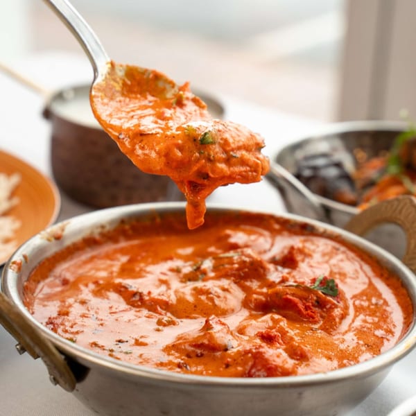A spoonful of vibrant orange Indian curry with chicken, dripping into a metal bowl.
