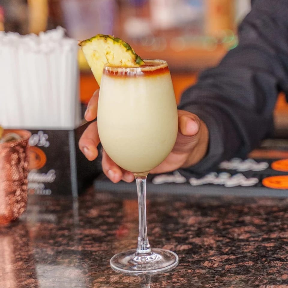 A hand holding a white cocktail with a pineapple slice garnish on a dark speckled bar.