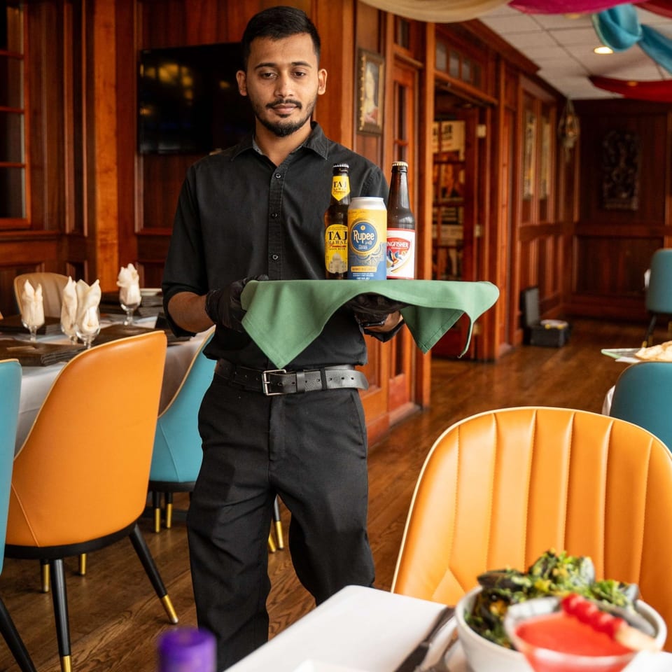 A Curry Club waiter in black uniform holds a tray with Indian beers and a can of Rupee.