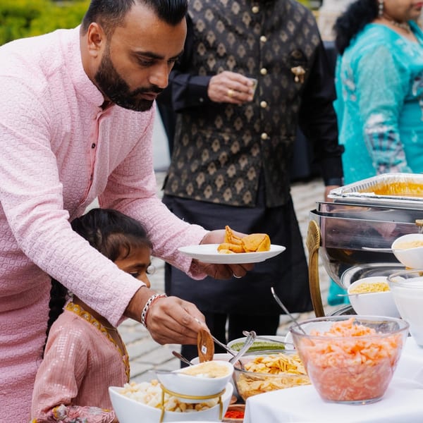 A man in a pink kurta serves samosas to a child at an Indian food buffet.