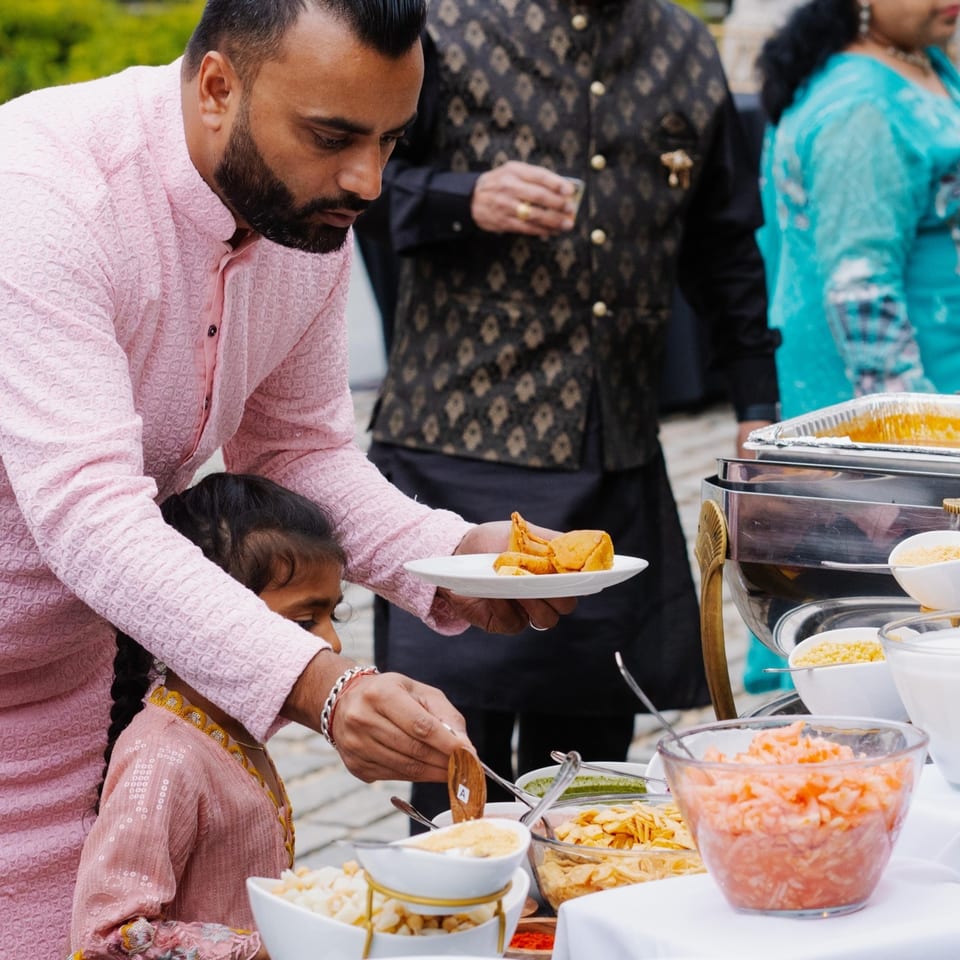 A man in a pink kurta serves samosas to a child at an Indian food buffet.