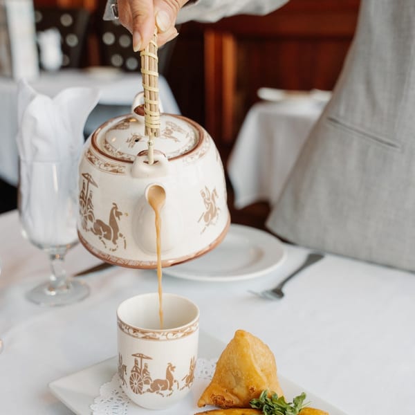 A hand pours chai from an ornate teapot into a matching cup, next to samosas at Curry Club.