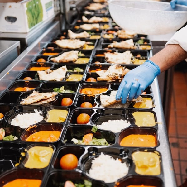 Gloved hands preparing multiple Indian meal prep containers with rice, curry, naan, and gulab jamun.