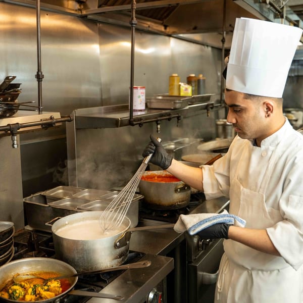 Curry Club chef in a white uniform and black gloves whisking a pot of Indian food.