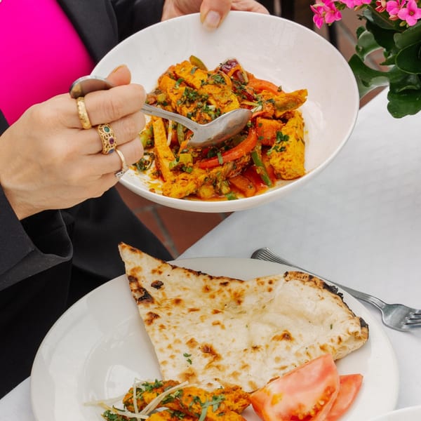 Hands serving Indian curry from a white bowl onto a plate with naan bread and tomato slices.