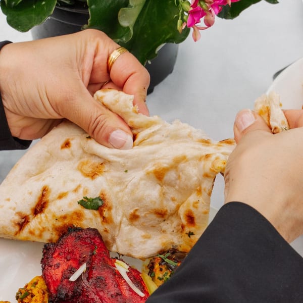 Hands tearing a piece of naan bread from a plate with Indian tandoori chicken and other dishes.