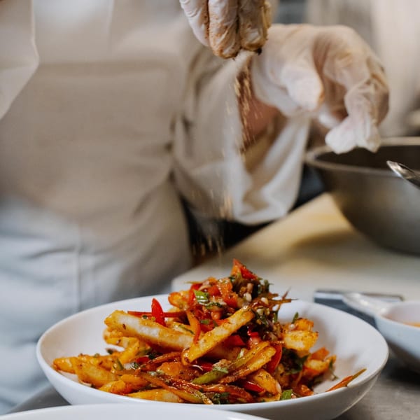 Chef in gloves sprinkling spices over a white bowl of chili paneer fries at Curry Club.
