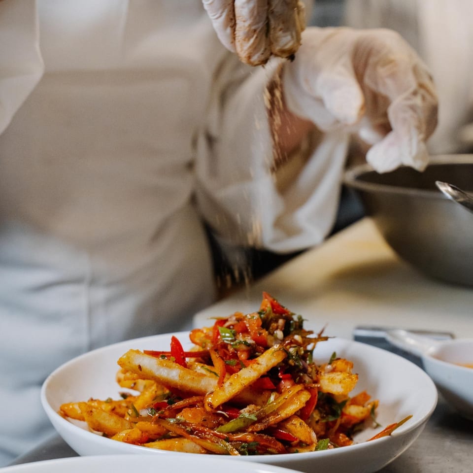 Chef in gloves sprinkling spices over a white bowl of chili paneer fries at Curry Club.