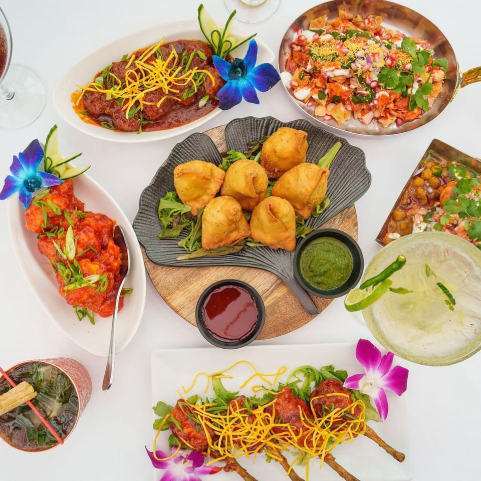 Overhead view of various Indian dishes including samosas, curries, and cocktails on a white table.