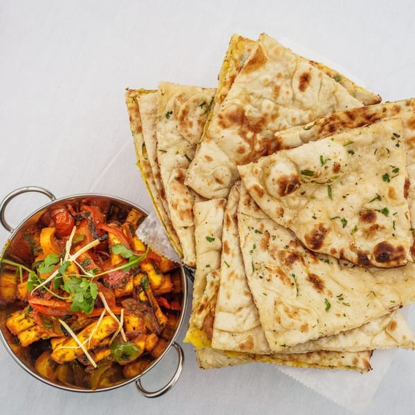 Indian paneer curry in a metal bowl next to a stack of naan bread on a white tablecloth.
