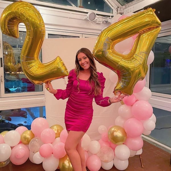 A smiling woman in a pink dress holds large gold '24' balloons, surrounded by pink and white balloons.