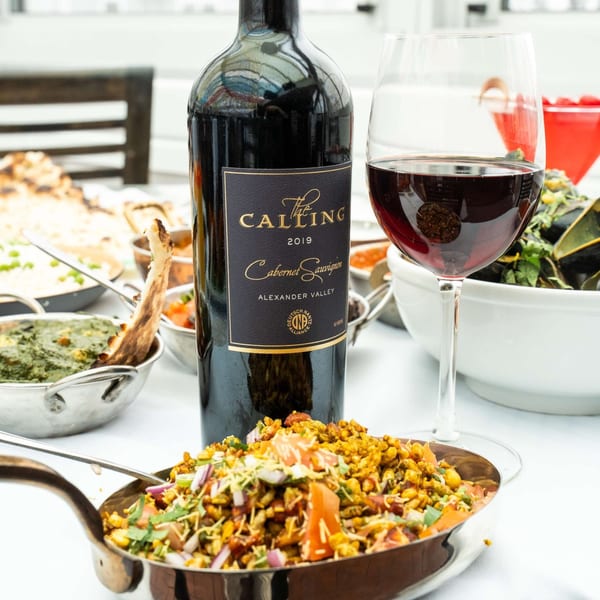 A bottle of "The Calling" Cabernet Sauvignon and a glass of red wine next to Indian dishes.