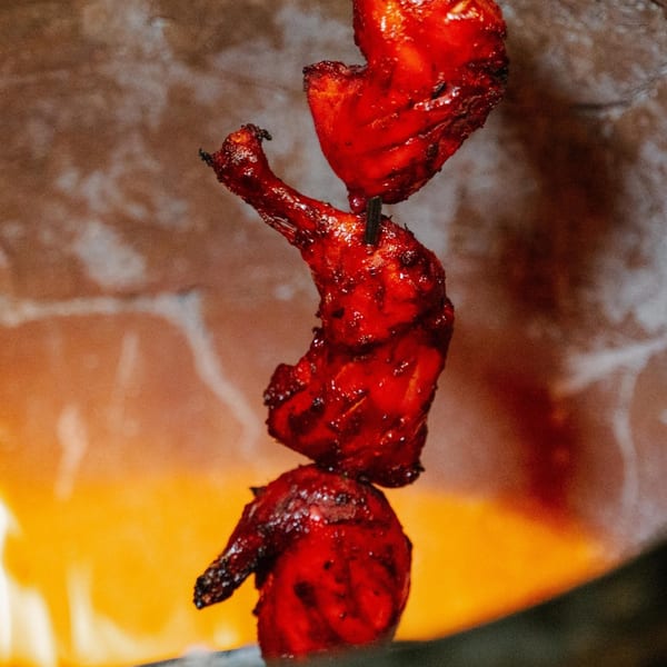 Three pieces of red tandoori chicken on a skewer cooking over a fiery orange flame.