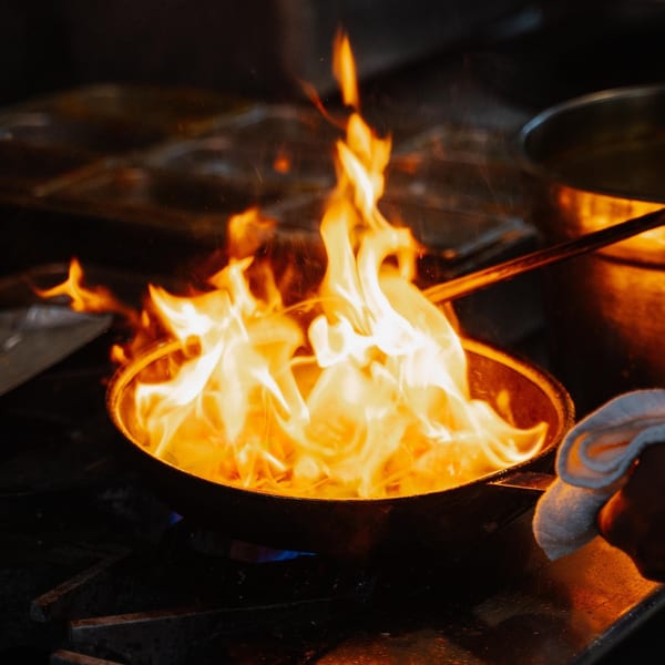 Chef flambéing Indian cuisine in a pan with large flames in a dark kitchen.