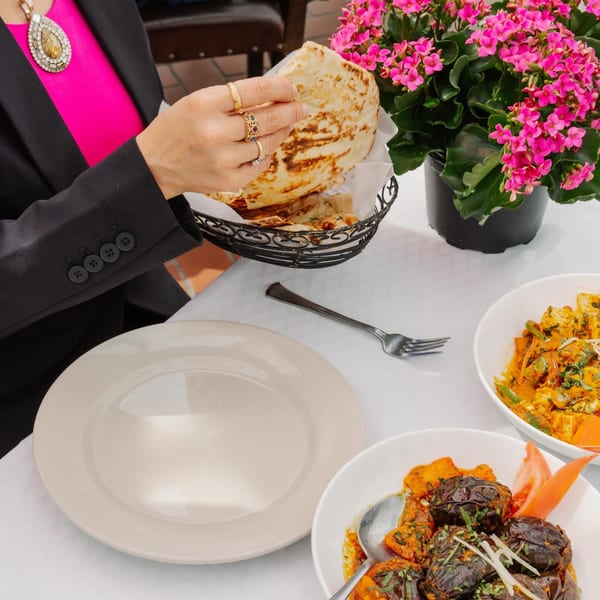 A person holding naan bread from a basket, with Indian dishes and a pink flower pot on a white table.
