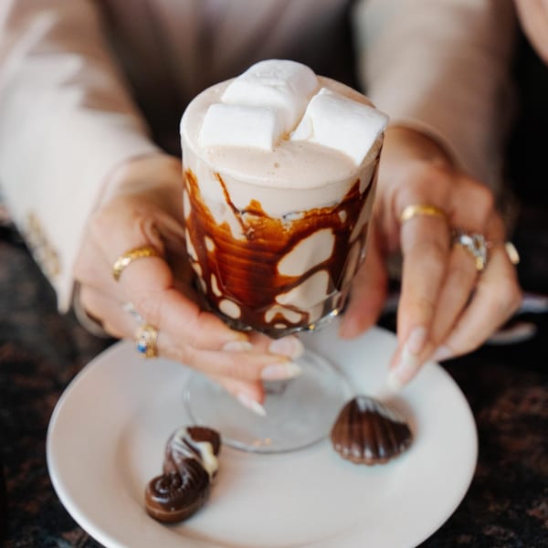Hands holding a glass of hot chocolate with marshmallows and chocolate syrup, with two chocolates on a plate.