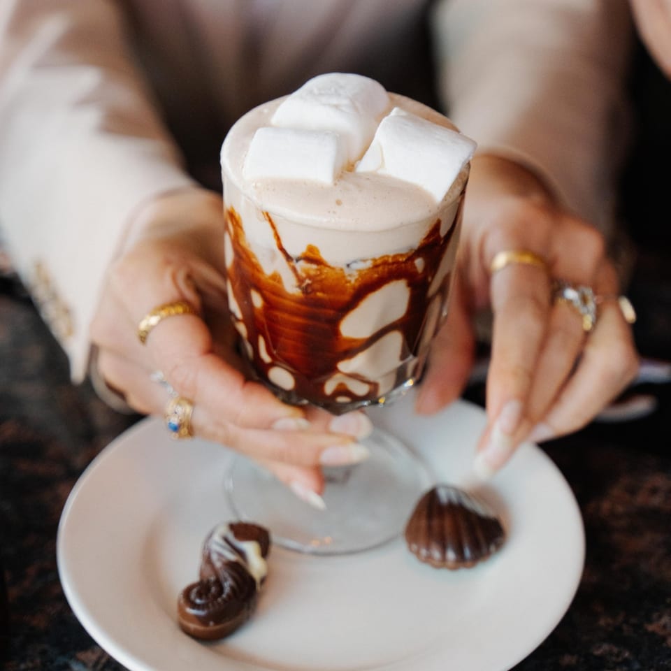 Hands holding a glass of hot chocolate with marshmallows and chocolate syrup, with two chocolates on a plate.