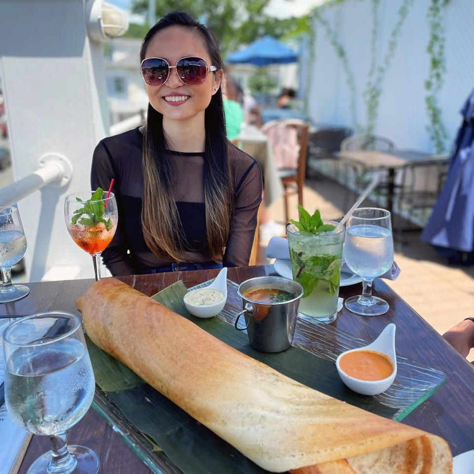 A smiling woman in sunglasses at Curry Club with a large dosa, chutneys, and cocktails on her table.