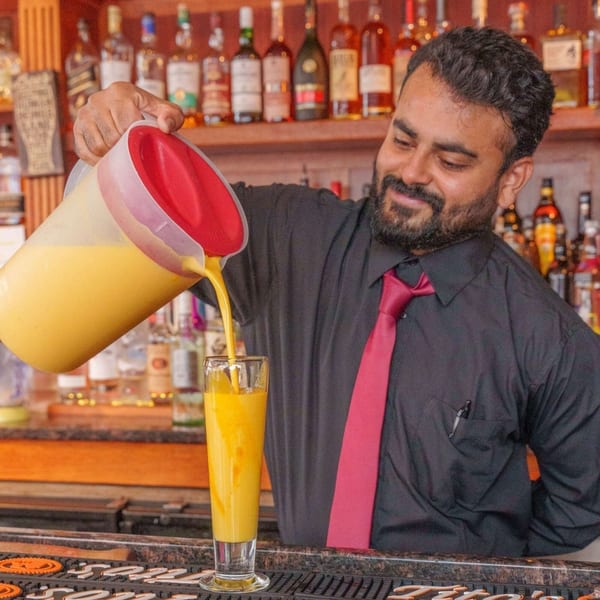 A bartender at Curry Club pours a yellow drink from a pitcher into a tall glass.