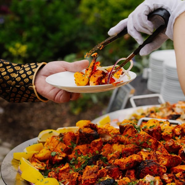 A gloved hand serves Indian tandoori chicken and shrimp from a large platter onto a plate.