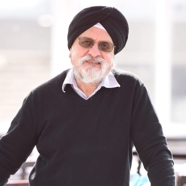An older man with a white beard and black turban smiles, wearing sunglasses and a black sweater.