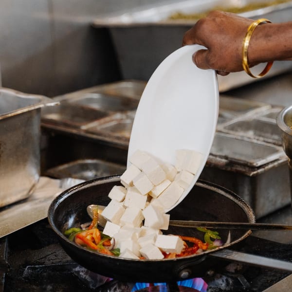 A chef's hand with a gold bracelet adds cubed paneer from a white plate into a pan of vegetables.
