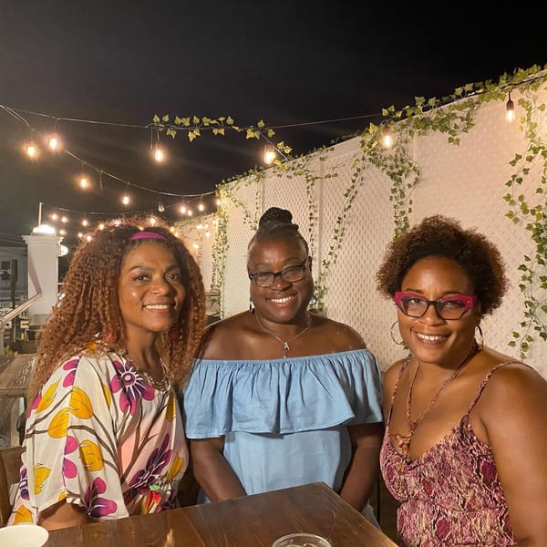 Three smiling women seated at a table on an outdoor patio with string lights and ivy.