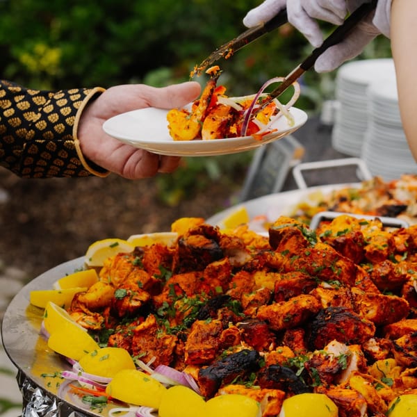 A server in white gloves uses tongs to serve Indian tandoori chicken from a large platter.