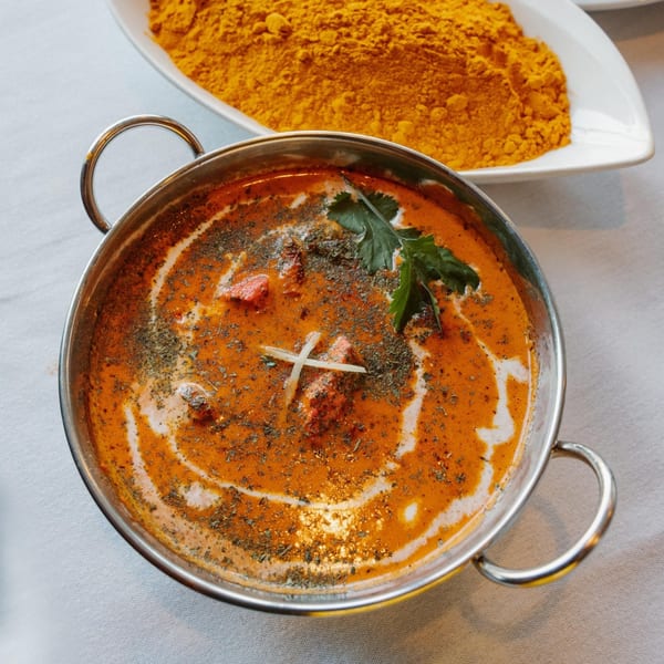 A metal bowl of Indian butter chicken with cream swirls, herbs, and a side of turmeric powder.