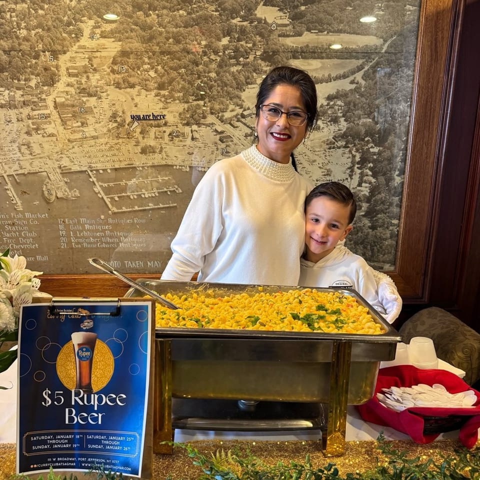 A woman and child smile behind a buffet of Indian macaroni and cheese, with a "$5 Rupee Beer" sign.
