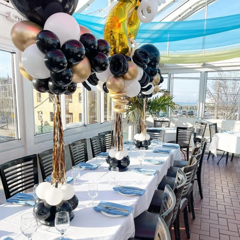 Decorated dining room at Curry Club with long tables, black and gold balloons, and a view of the water.