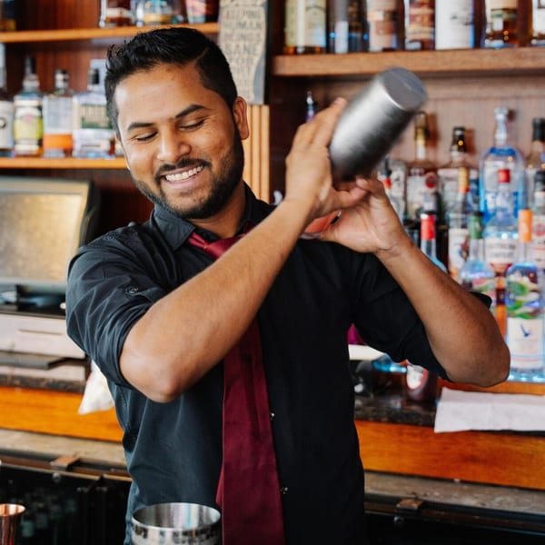 A smiling bartender at Curry Club shakes a cocktail shaker with bottles of liquor in the background.