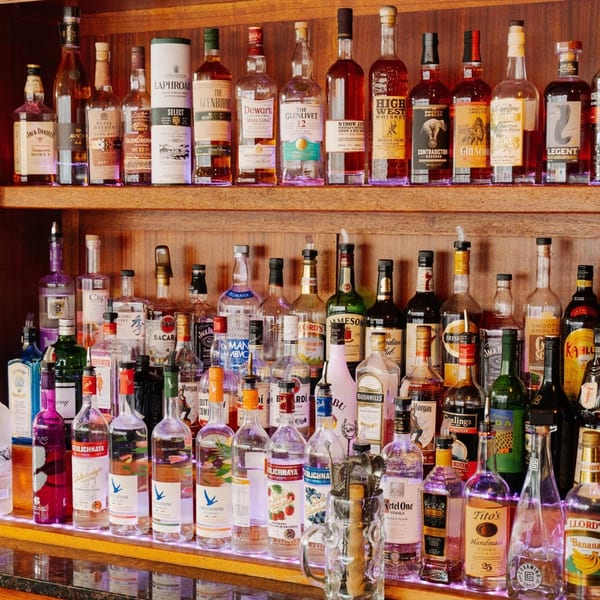 A well-stocked bar with two shelves full of various liquor bottles, illuminated by purple lights.