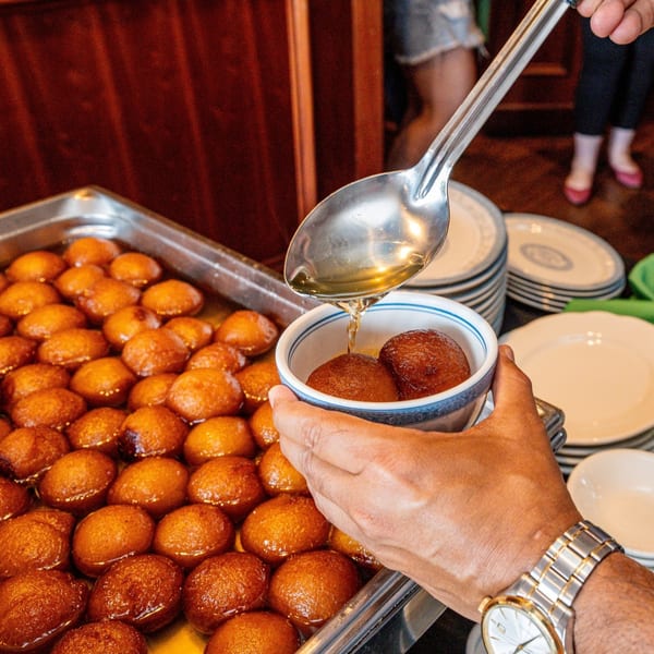 A hand serving Gulab Jamun, an Indian dessert, from a large tray into a small bowl.