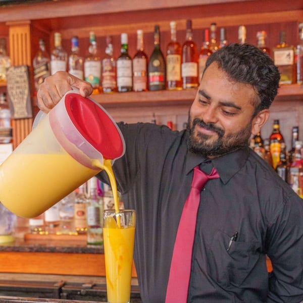 A smiling bartender pours a yellow drink from a pitcher into a tall glass at Curry Club.