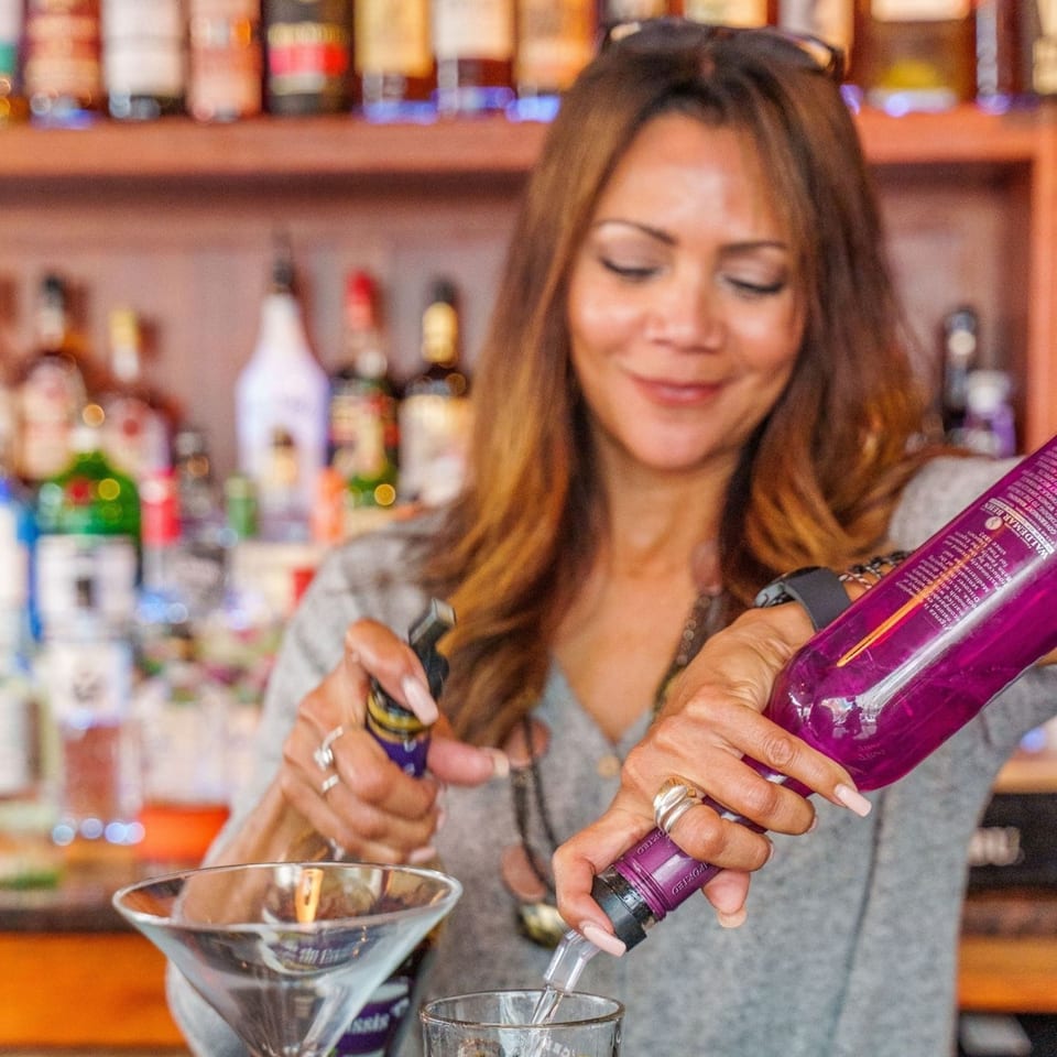 A smiling bartender pours a drink from a purple bottle into a glass at Curry Club.