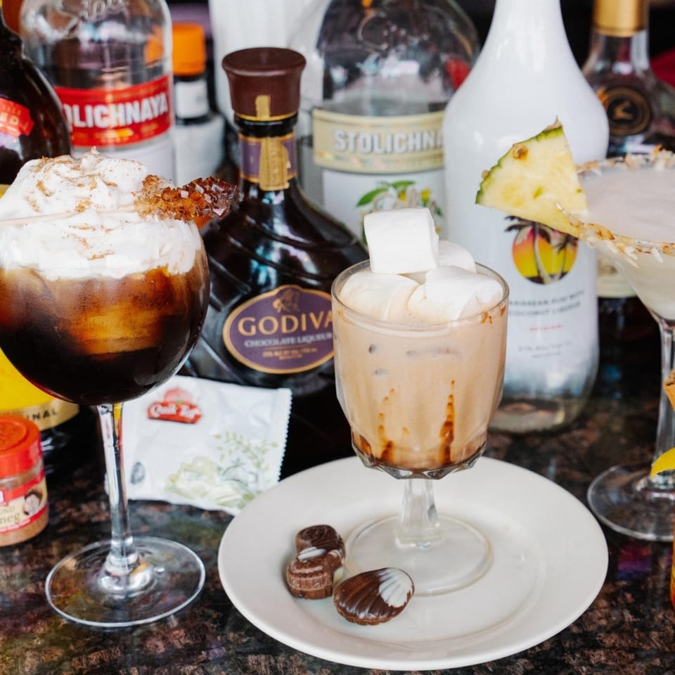 Two cocktails, one dark with whipped cream, one light with marshmallows, surrounded by liquor bottles.