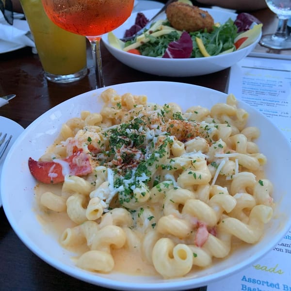 Creamy macaroni and cheese with lobster, parsley, and spices, alongside a salad and drinks.