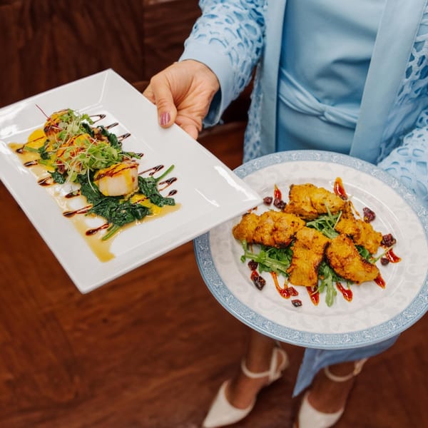 A server in a blue dress holds two plates of Indian cuisine, one with scallops, the other with chicken.