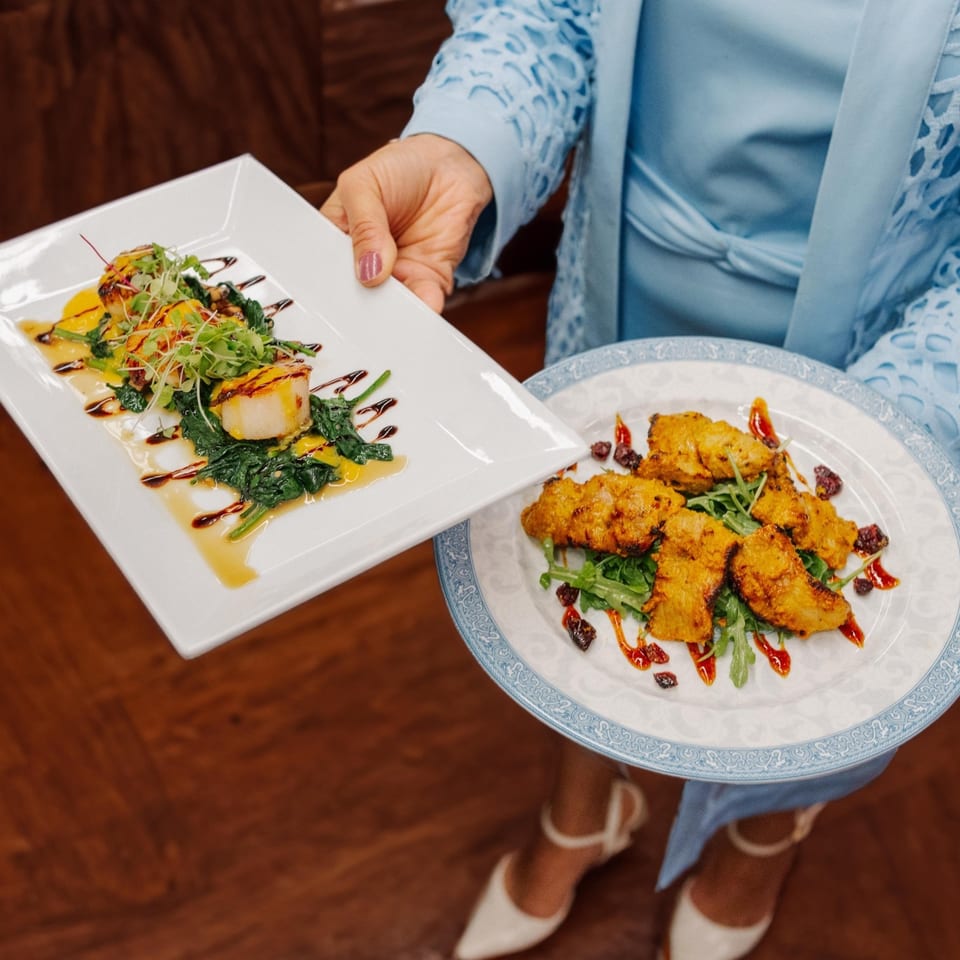 A server in a blue dress holds two plates of Indian cuisine, one with scallops, the other with chicken.