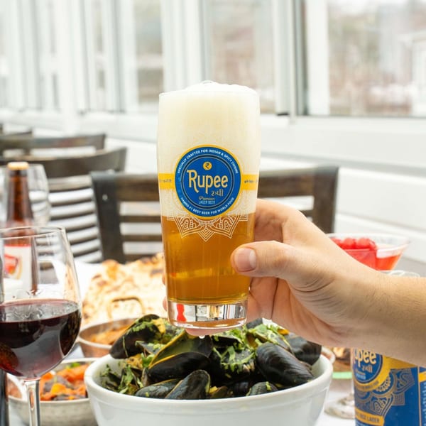 A hand holding a glass of Rupee Lager Beer above a bowl of mussels and other Indian dishes.