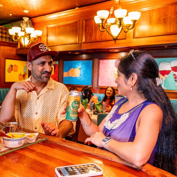 A man and woman at a bar, the woman holding a Rupee beer can, with colorful art in the background.